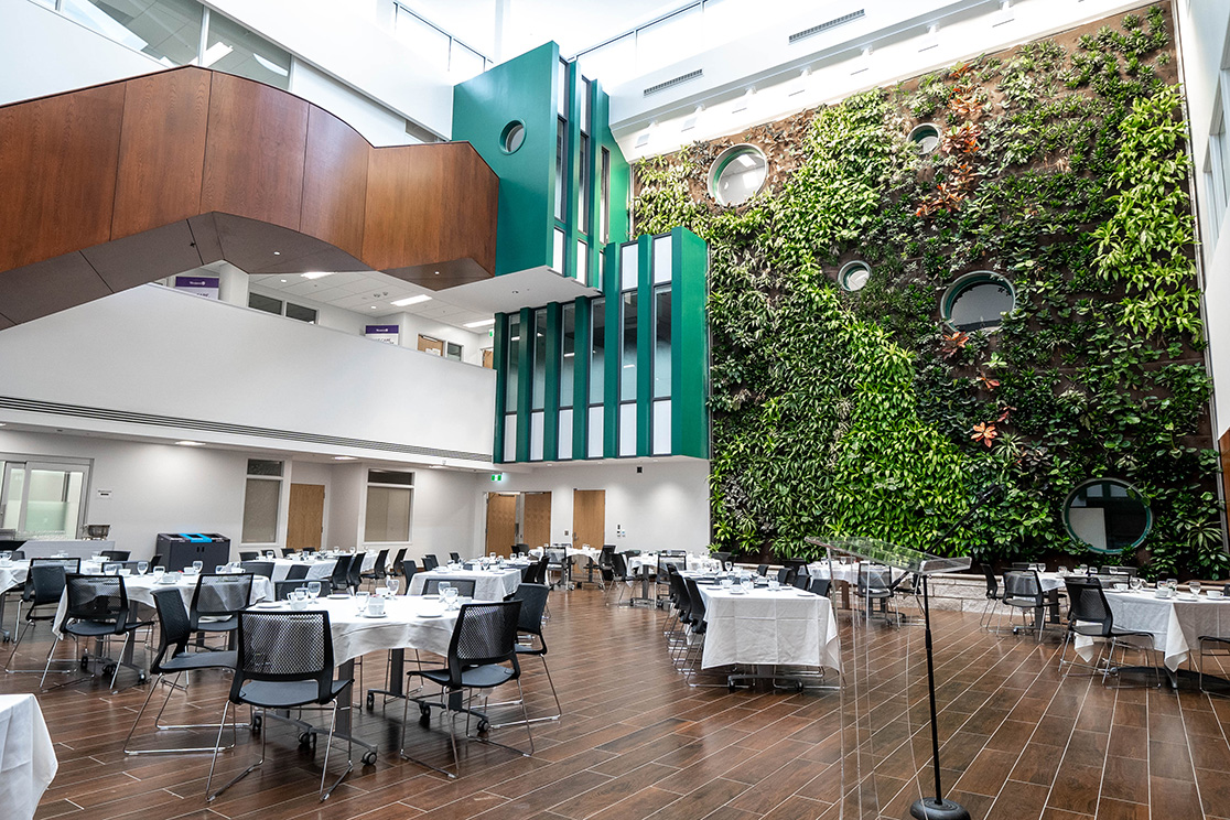 Speaker addressing seated guests at round tables in a modern atrium with a floor-to-ceiling living wall, wood flooring, and white walls. The space features natural light, green vertical garden, and contemporary architecture.